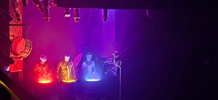 A stage, the only light coming from three large drums, one glowing with red paint, one yellow, one blue. Behind each drum stands a Blue Man, a bald, blue humanoid in simple black clothes.