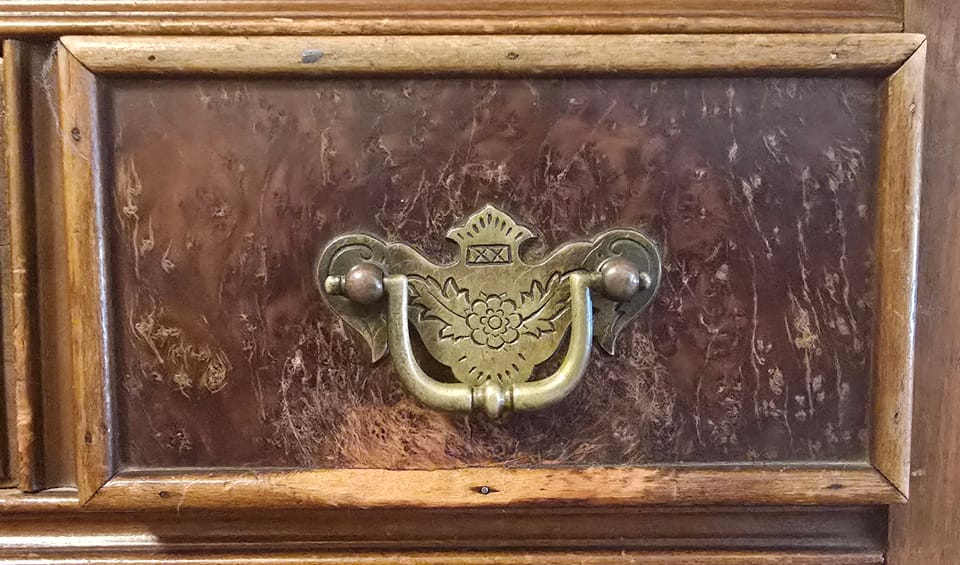 A close-up of the front of a drawer. Dark wood, engraved brass pull, deep scratches in the wood below.